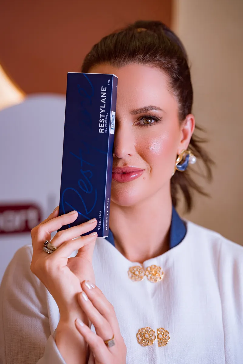Mulher elegante segurando caixa azul de preenchimento Restylane Galderma junto ao rosto, brincos dourados com pérola, blazer branco, fundo bege e terracota, fotografia editorial de luxo