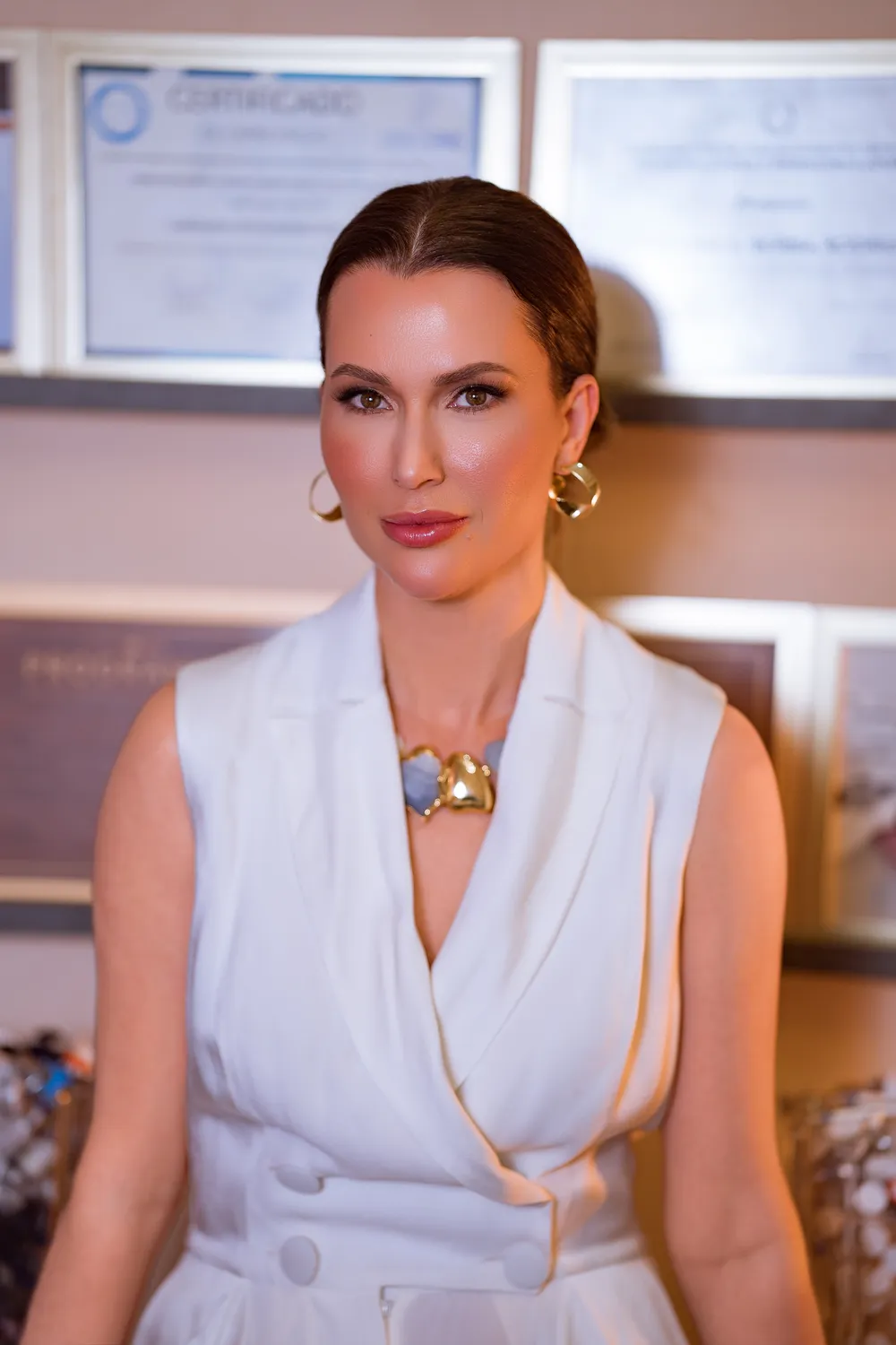 Retrato de mulher elegante de vestido branco com acessórios dourados, expressão serena e sofisticada, fundo de clínica com certificados emoldurados, iluminação editorial quente, fotografia de luxo