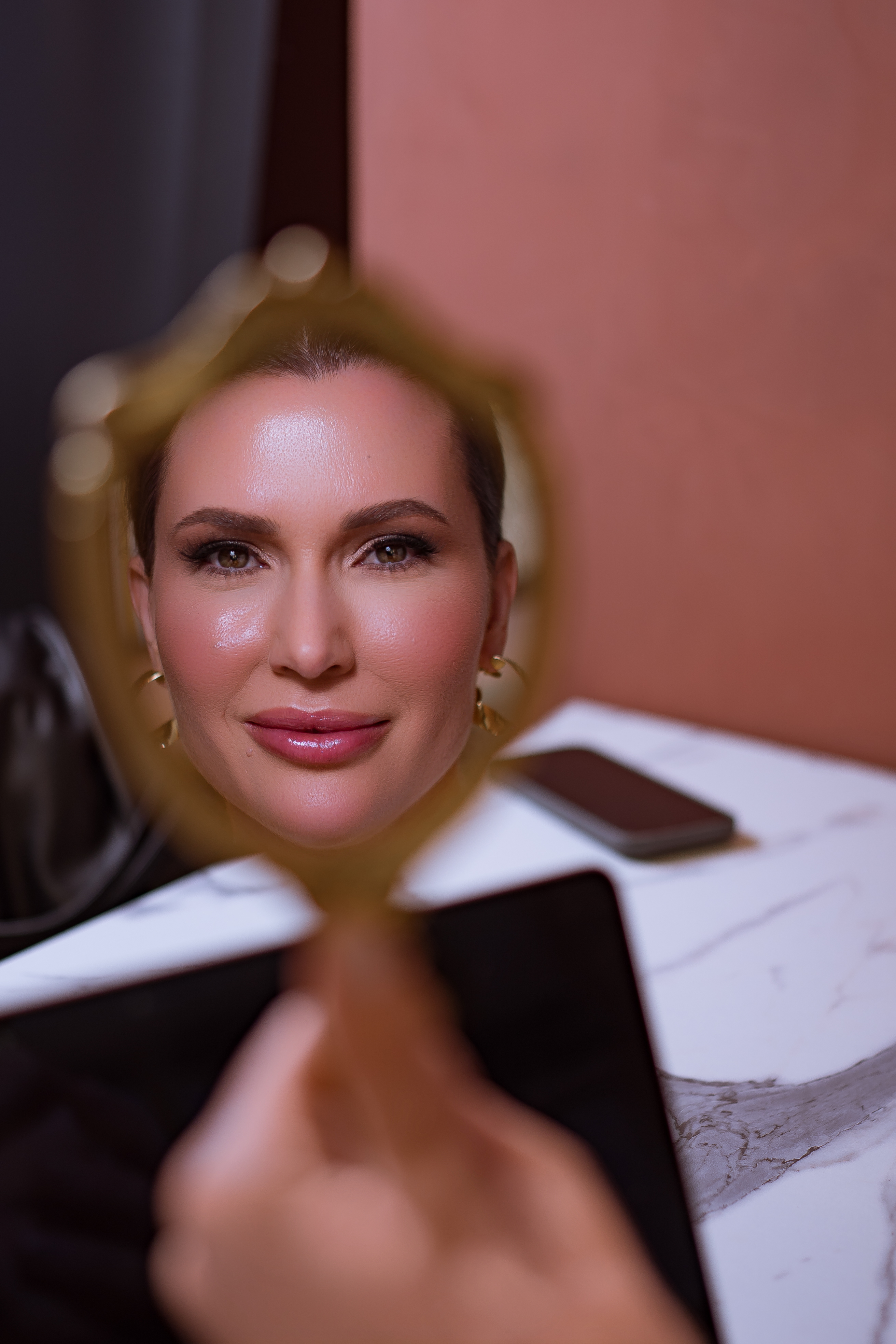 Retrato de mulher elegante refletida em espelho de mão dourado, pele luminosa e maquiagem sofisticada, fundo em tons terracota e mármore, iluminação quente editorial de luxo