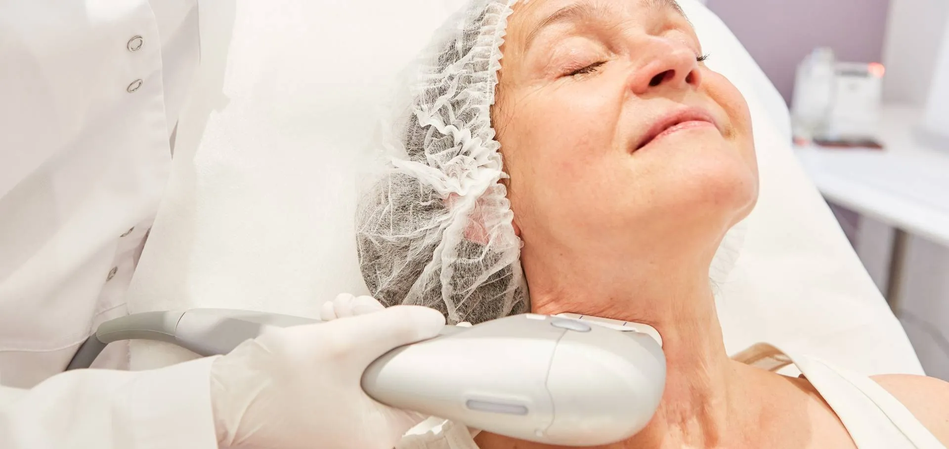 Mulher madura recebendo aplicação de ultrassom microfocado no pescoço em clínica de luxo, aplicador conduzido por profissional com luva, pele luminosa, ambiente claro em tons bege e branco, fotografia beauty premium